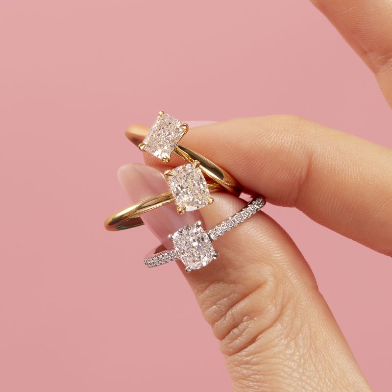 Three solitaire diamond engagement rings held by a model, featuring an elongated radiant cut diamond in 18ct yellow gold, an elongated cushion cut diamond with a hidden halo in 18ct yellow gold, and a